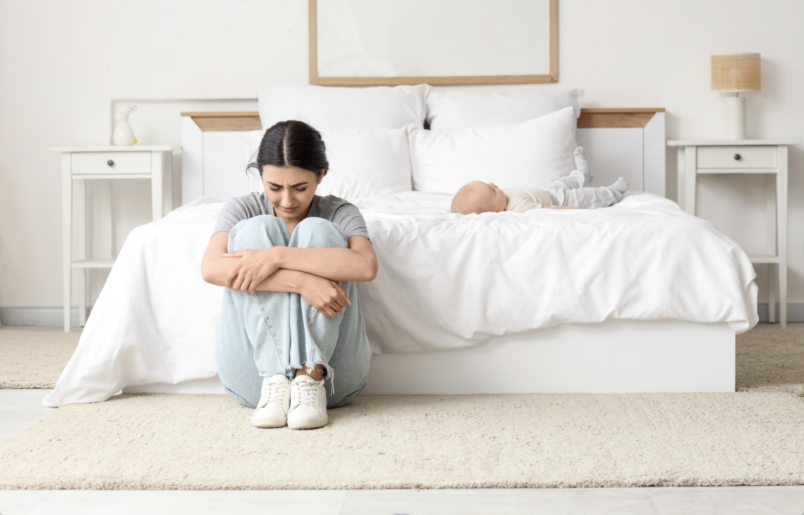 New mother sitting hugging knees on bedroom floor struggling with postpartum depression while baby lies on bed